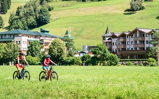 Zwei Fahrradfahrende in der grünen Natur mit Gebäuden im Hintergrund.