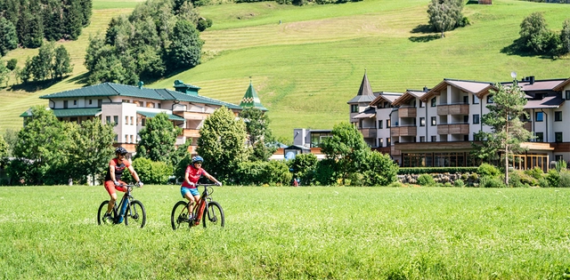 SporthotelSillian_Hero_C-GertPerauer Zwei Fahrradfahrende in der grünen Natur mit Gebäuden im Hintergrund.