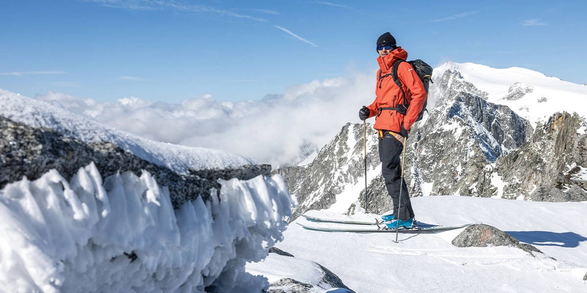 Person auf Ski mit Blick in die Kamera vor verschneiter Bergkulisse.