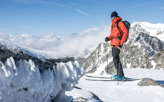 Person auf Ski mit Blick in die Kamera vor verschneiter Bergkulisse.