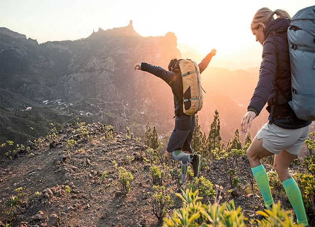 Zwei Wanderer hüpfen bei Sonnenuntergang in steinigen Gelände den Berg hinunter.