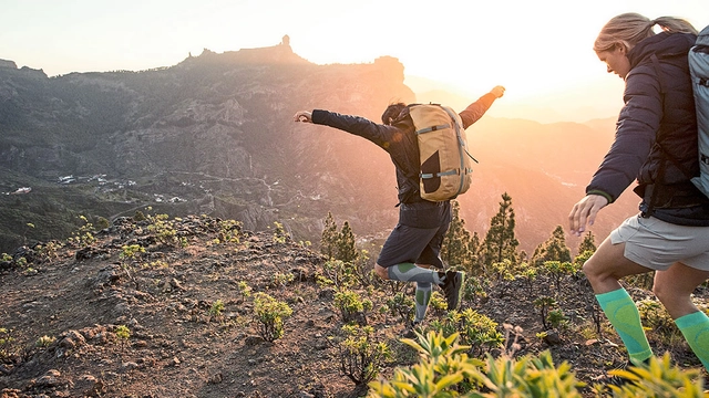 Zwei Wanderer hüpfen bei Sonnenuntergang in steinigen Gelände den Berg hinunter.