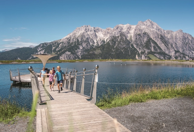 Eine Familie erkundet das Ufer eines Sees, auf einem Steg mit Bergpanorama im Hintergrund