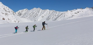 Vier Skitourengeher vor schneebedeckter Bergkulisse bei blauem Himmel.