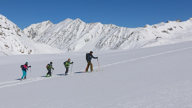 Vier Skitourengeher vor schneebedeckter Bergkulisse bei blauem Himmel.