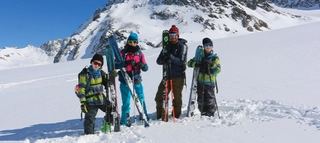 Vier Personen vor verschneiten Bergen mit Ski im Schnee stehend.