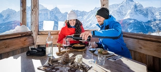 Zwei Personen beim Essen in einem Gebäude im Skigebiet 3 Zinnen mit Bergen im Hintergrund.
