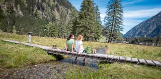 Zwei Frauen sitzen auf einer kleine Brücke über einem Wiesenbach und lachen. Im Hintergrund ist ein kleiner See.