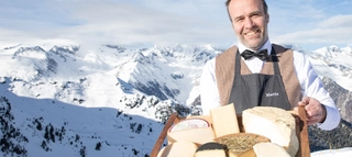 Eine Person mit Tablett mit Käse vor verschneiten Bergen in Südtirol.