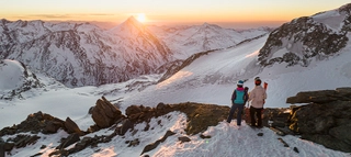 Zwei Personen mit Ski vor verschneiten Bergen mit Sonne am Horizont im Saastal.