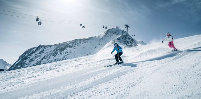 Zwei Skifahrer auf der Piste mit Gondelbahn im Hintergrund.