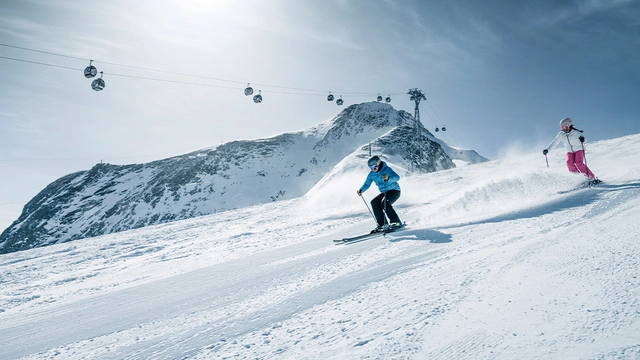 Zwei Skifahrer auf der Piste mit Gondelbahn im Hintergrund.