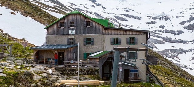 Aufnahme einer Berghütte mit Schnee im Hintergrund.