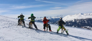 Personen auf Snowbikes sitzend vor verschneitem Bergpanorama.