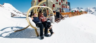 Zwei Personen vor einem Gebäude und schneebedeckten Bergen in der Ferienregion Saas-Fee/Saastal.