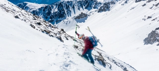 Person, die mit an Rucksack befestigten Ski einen verschneiten Berg hinaufklettert.