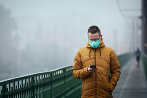 Lonely man with face mask using phone during walk on bridge against city in mysterious fog. Gloomy weather in Prague, Czech Republic.