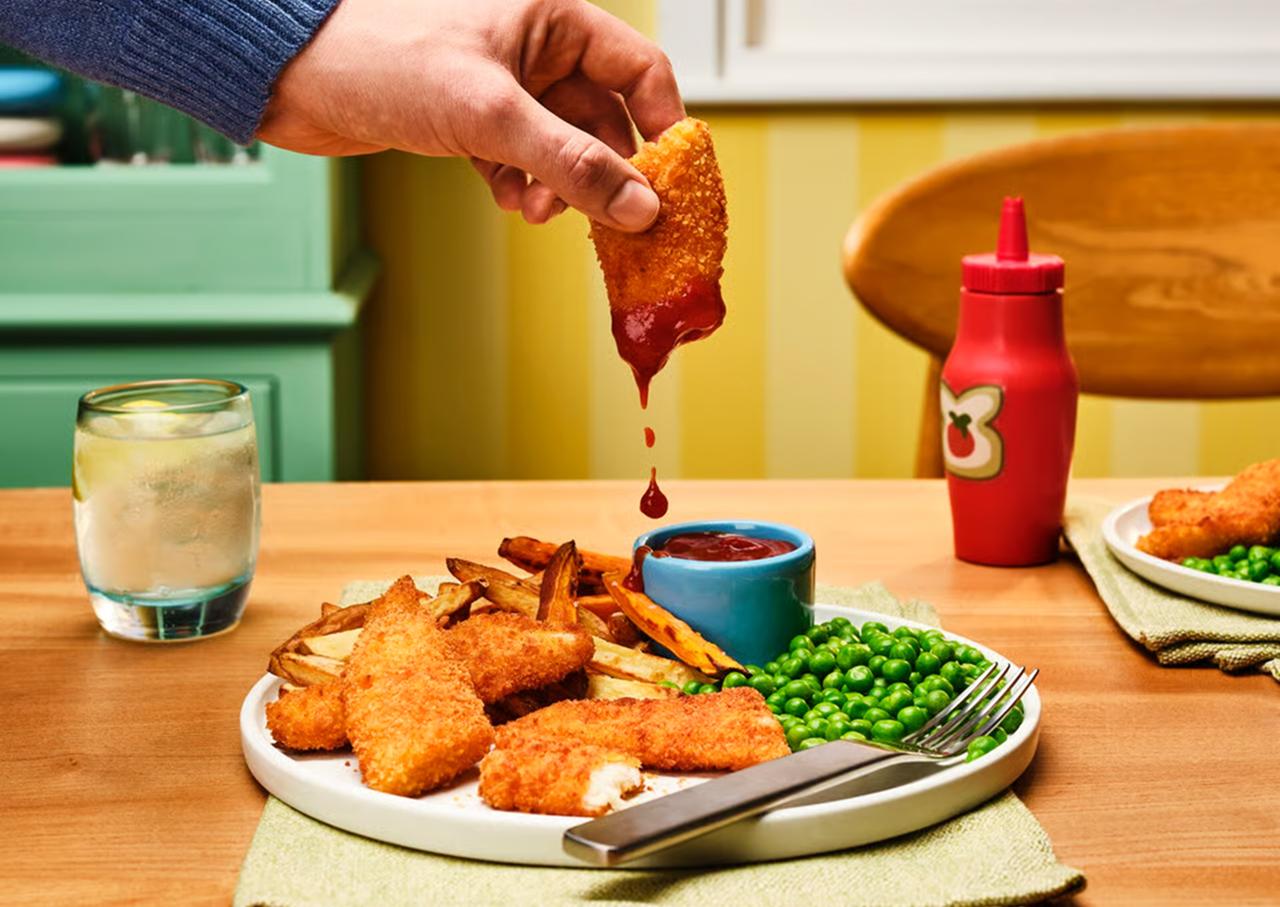 A plate of Gousto's fish, chips and peas, with a fish finger being dipped into tomato ketchup.
