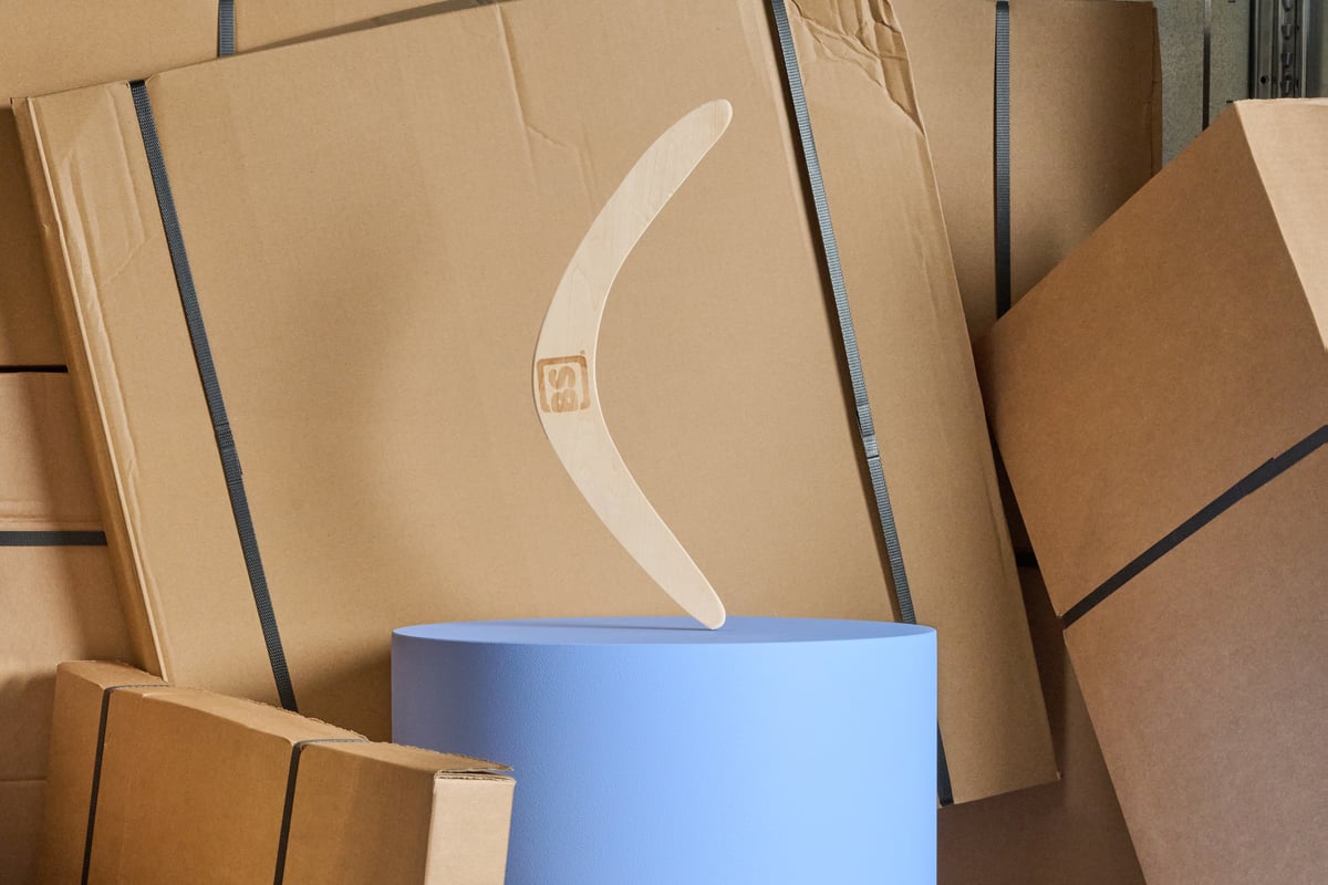 A wooden boomerang rests on a blue cylindrical stand, surrounded by large stacked cardboard boxes.