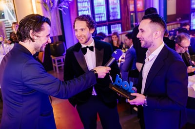 Een glimlachende man houdt een microfoon vast en interviewt twee mannen in pak tijdens een prijsuitreiking.