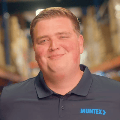 Een glimlachende persoon in een donker poloshirt met "MUNTEX"-logo staat in een magazijngang met planken met dozen. De sfeer is warm en uitnodigend.