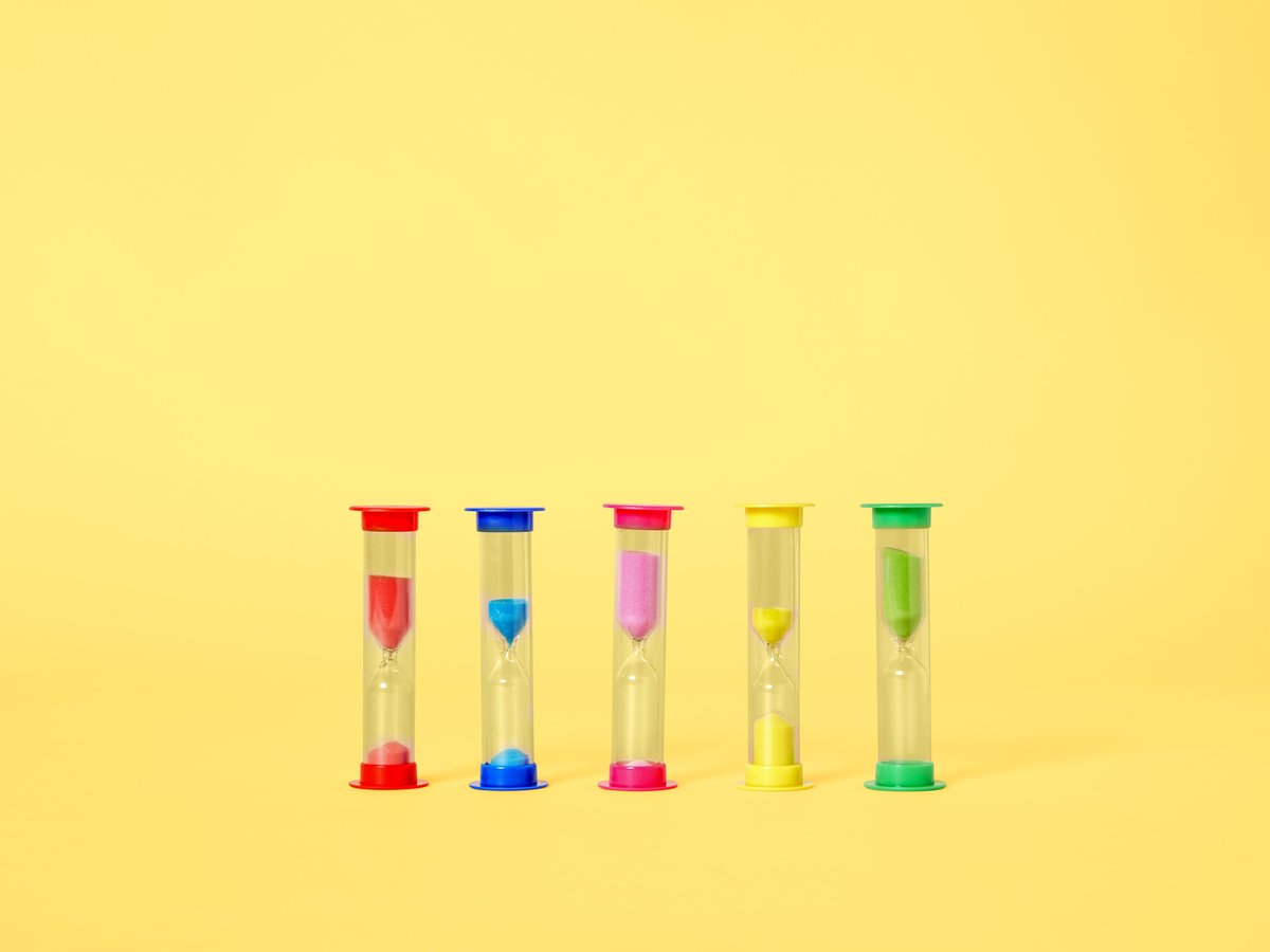 Vijf kleurrijke zandlopers op een rij op een gele achtergrond. Elke zandloper heeft een andere kleur zand: rood, blauw, roze, geel en groen.