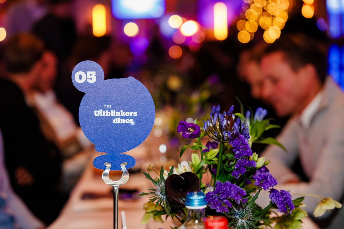 Elegant dining table decorated with purple flowers and a blue plate with the text ‘The Achievers Dinner 05’. In the background, blurred guests are having a warm conversation.