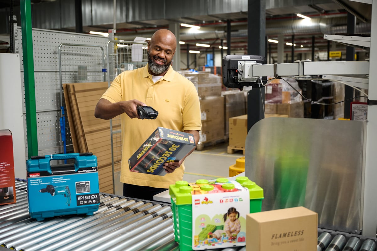 Een man in een geel shirt gebruikt een barcodescanner op een kleurrijke speelgoeddoos in een magazijn.