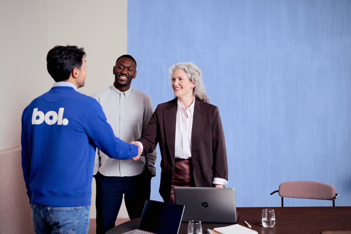 Drie mensen tijdens een overleg. Twee geven elkaar een hand. Eén persoon draagt een blauwe trui met het bol-logo op de rug.