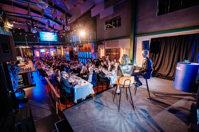 Een levendig, formeel diner in een ruime zaal met een hoog plafond. Twee sprekers op het podium staan tegenover een groot, zittend publiek aan met kaarsen verlichte tafels.