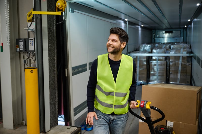 Een man in een gele veiligheidsvest gebruikt een palletwagen om dozen te verplaatsen in een magazijn, terwijl op de achtergrond een met pallets beladen vrachtwagen zichtbaar is.