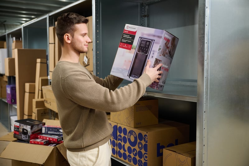 A man in a beige sweater places a box on a shelf in a storage room, surrounded by other boxes.