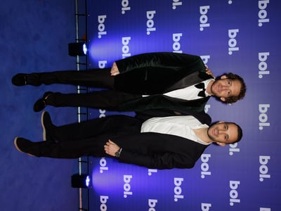 Twee mannen in pak poseren op een blauw tapijt tegen een achtergrond met herhaaldelijk ‘bol’.