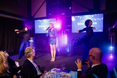 Een vrouw in een sprankelende blauwe jurk zingt op het podium, geflankeerd door twee dansers in bijpassende outfits. 