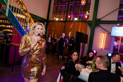 Een vrouw in een gouden jurk houdt een microfoon vast en glimlacht tijdens een feestelijk evenement.