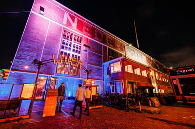 Een levendig nachtelijk tafereel van een bakstenen gebouw dat verlicht wordt door roze en blauwe lichten. Boven de ingang hangt het neonbord ‘IJVER’.