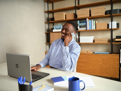 Smiling businessman talks on a smartphone at his desk beside an open laptop, blue mug, notebook, and office shelves; relaxed mood.
