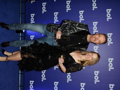 A couple smiles on a blue carpet at an event. The man wears a patterned blazer and jeans, holding a champagne glass. The woman wears a black lace dress.