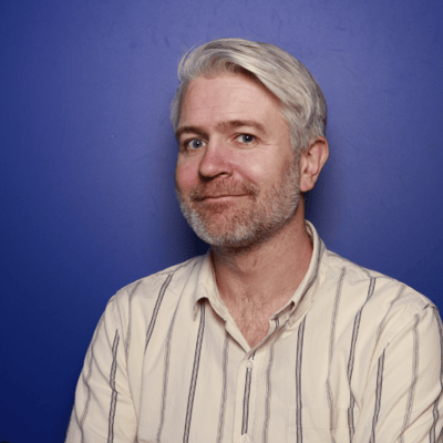 Middle-aged man with gray hair and a beard, wearing a striped shirt, smiles slightly against a solid blue background.