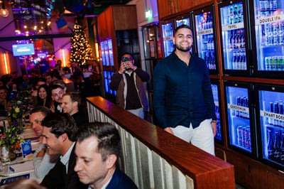 Een glimlachende man in een donker shirt staat naast koelkasten vol drankjes, naast een feestelijke eetruimte. 