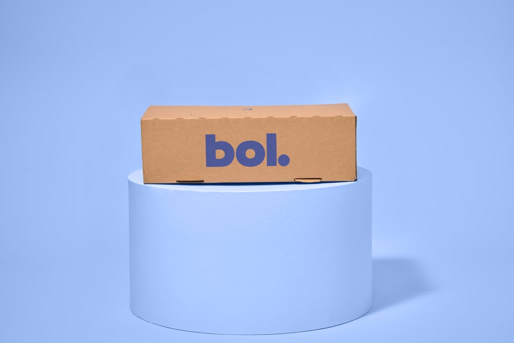 Een bruine kartonnen doos met ‘bol.’ in blauw staat op een cilindervormig wit voetstuk tegen een lichtblauwe achtergrond.