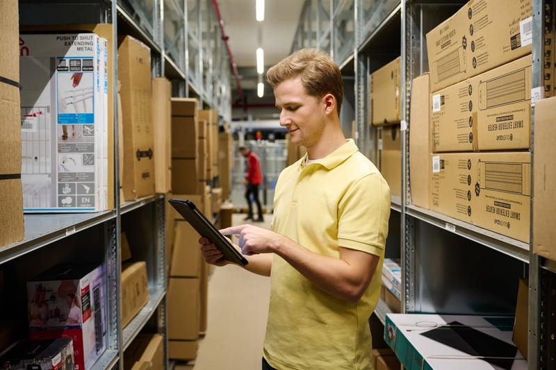 A man in a yellow shirt uses a tablet in a warehouse aisle with large, stacked boxes.