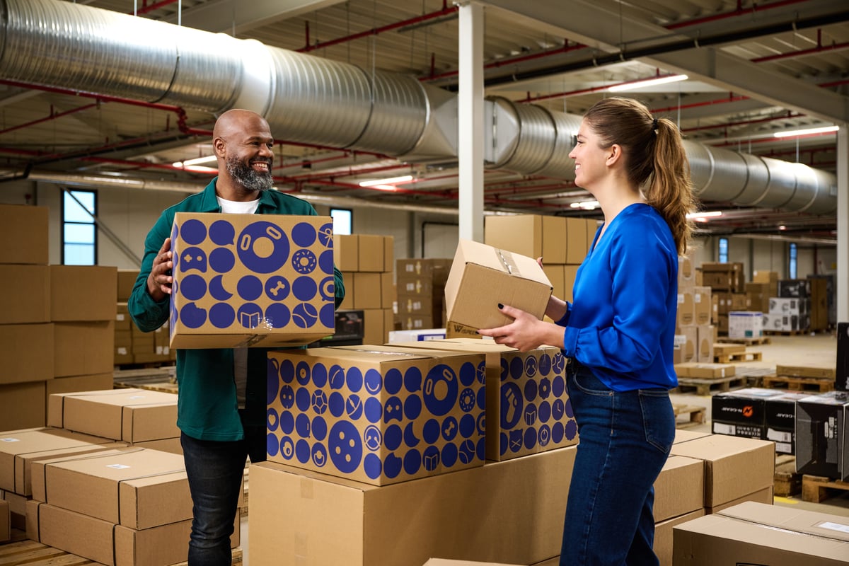 Een glimlachende man en vrouw staan in een magazijn vol dozen en wisselen pakketten uit.