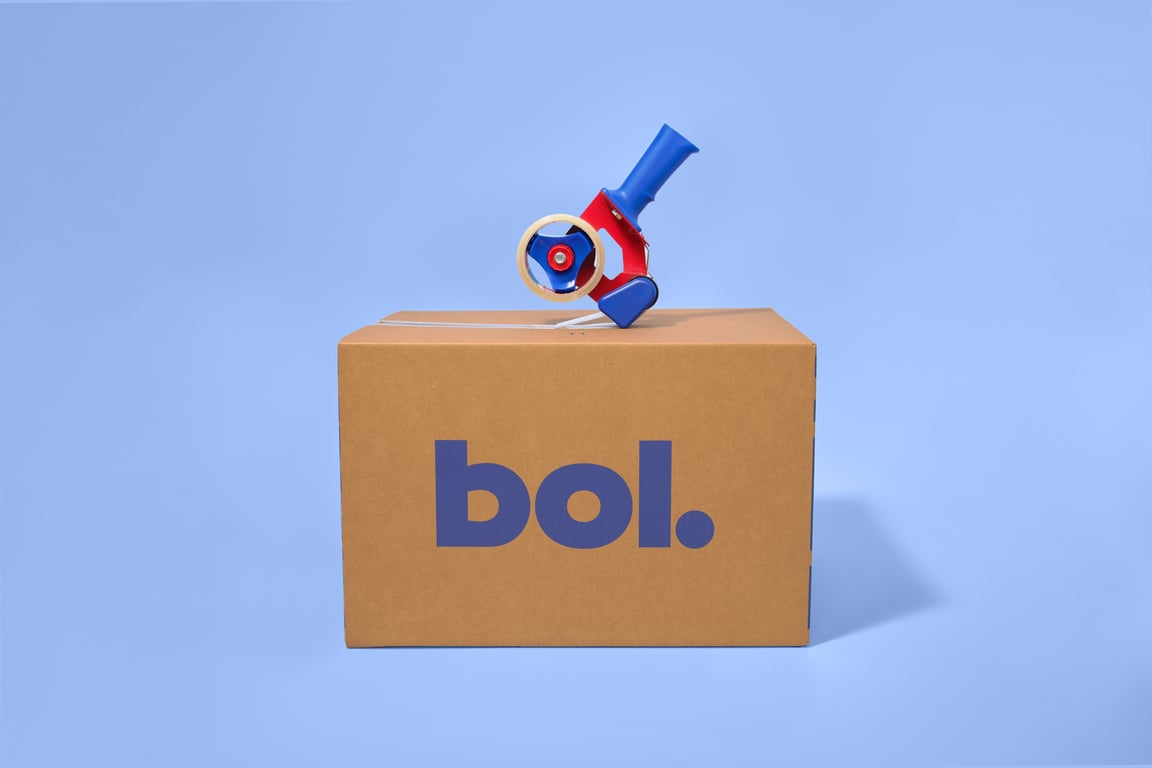 Cardboard box with "bol." logo in purple, set against a blue background. A red, blue, and white tape dispenser rests on top, evoking a neat, organized feel.