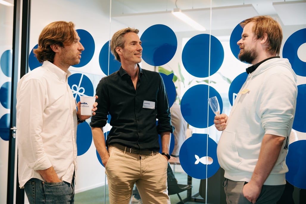 Three men are casually talking in an office with blue dotted glass