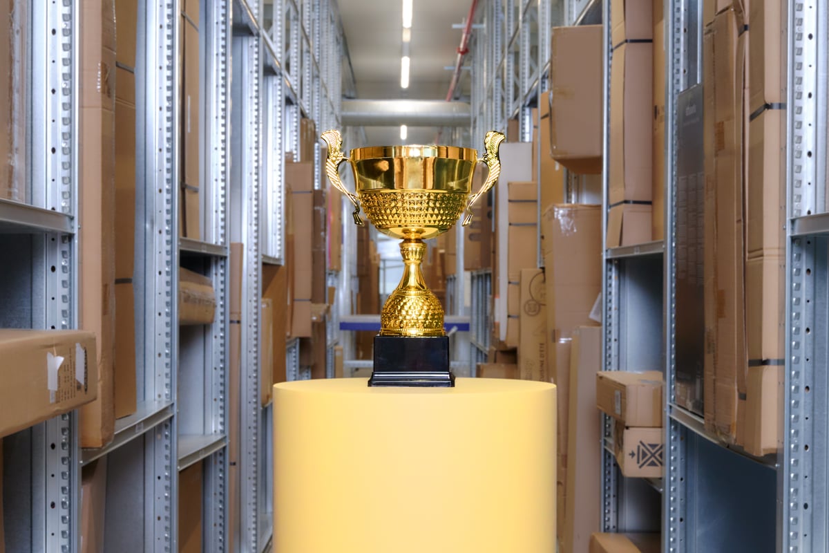 A shiny golden trophy stands on a yellow pedestal in the middle of a warehouse aisle full of brown boxes on metal shelves.