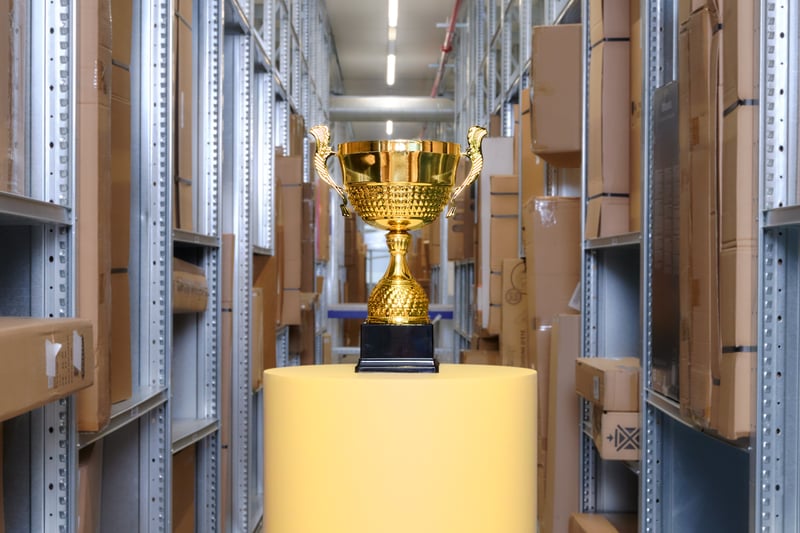 Een gouden trofee op een geel voetstuk staat centraal in een gang met hoge schappen vol kartonnen dozen, wat een gevoel van prestatie en focus uitstraalt.