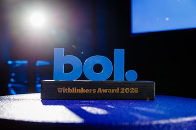 Blauw verlichte trofee met ‘bol.’ bovenop en ‘Uitblinkers Award 2025’ gegraveerd, geplaatst op een tafel met felle spotlights en een subtiel achtergrondscherm.