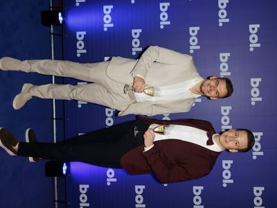 Two men in formal attire stand smiling on a blue carpet, holding champagne glasses. Behind them is a branded "bol." backdrop, creating a celebratory mood.