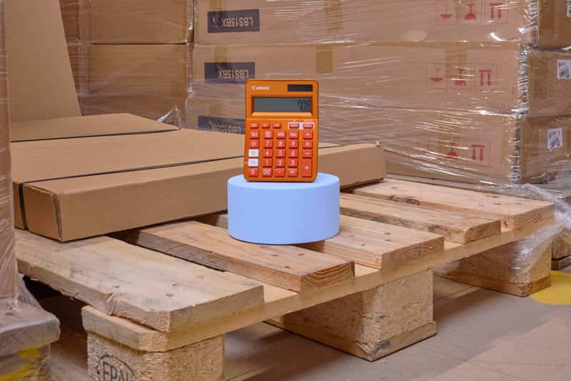 A warehouse scene with stacked cardboard boxes on wooden pallets. On top of these is a blue cylinder with an orange calculator on it.